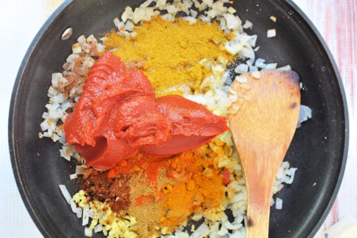 Chopped onions, spices, and tomato paste in a skillet with a wooden spatula&mdash;perfect for starting a rich Chicken Makhani before transferring to the slow cooker.