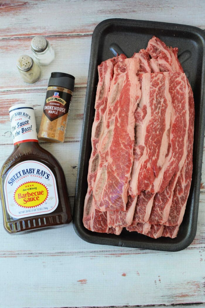 Raw beef riblets on a tray with barbecue sauce, smokehouse maple seasoning, salt, and pepper shakers nearby—perfect for your next beef riblets recipe.