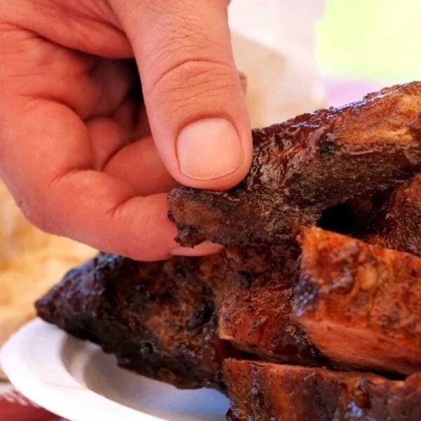 A hand picking up a barbecued rib from a plate on a checkered tablecloth, showcasing the delicious results of learning how to grill ribs on gas grill.