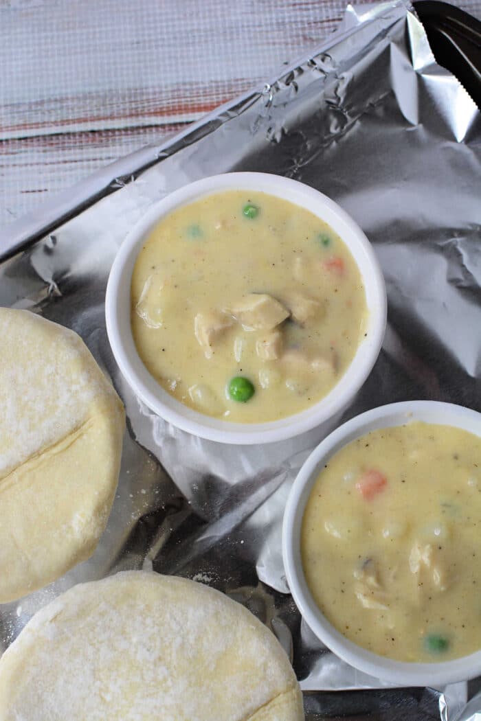 Two ramekins filled with creamy Chicken Pot Pie filling, next to uncooked pastry rounds on a foil-lined tray—a cozy recipe reminiscent of KFC’s classic comfort food.