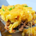 A spatula lifts a cheesy serving of meatloaf mac and cheese casserole from a baking dish, topped with chives.