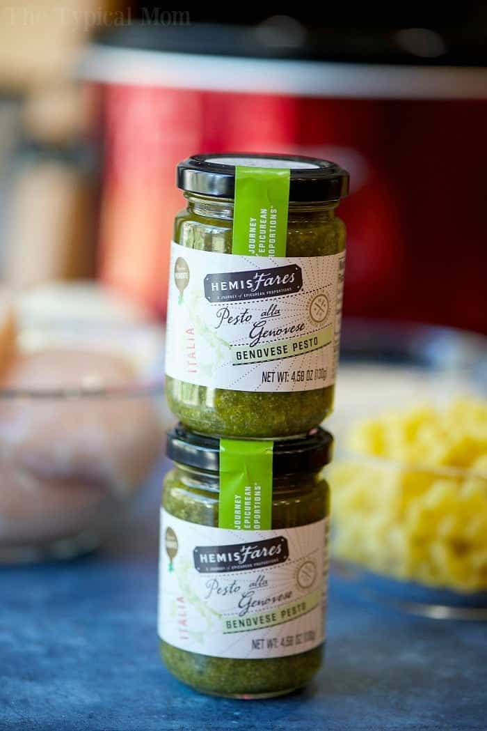 This easy crockpot pesto chicken alfredo casserole is a hit in my house! Cheesy noodles with moist pieces of pesto chicken together makes a complete meal. Two jars of HemisFares Genovese pesto stacked on a table with blurred ingredients in the background, perfect for adding bold flavor to your next crockpot pesto chicken alfredo casserole.