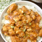 Bowl of rice topped with spicy honey Instant Pot chicken pieces, garnished with sesame seeds and sliced green onions.