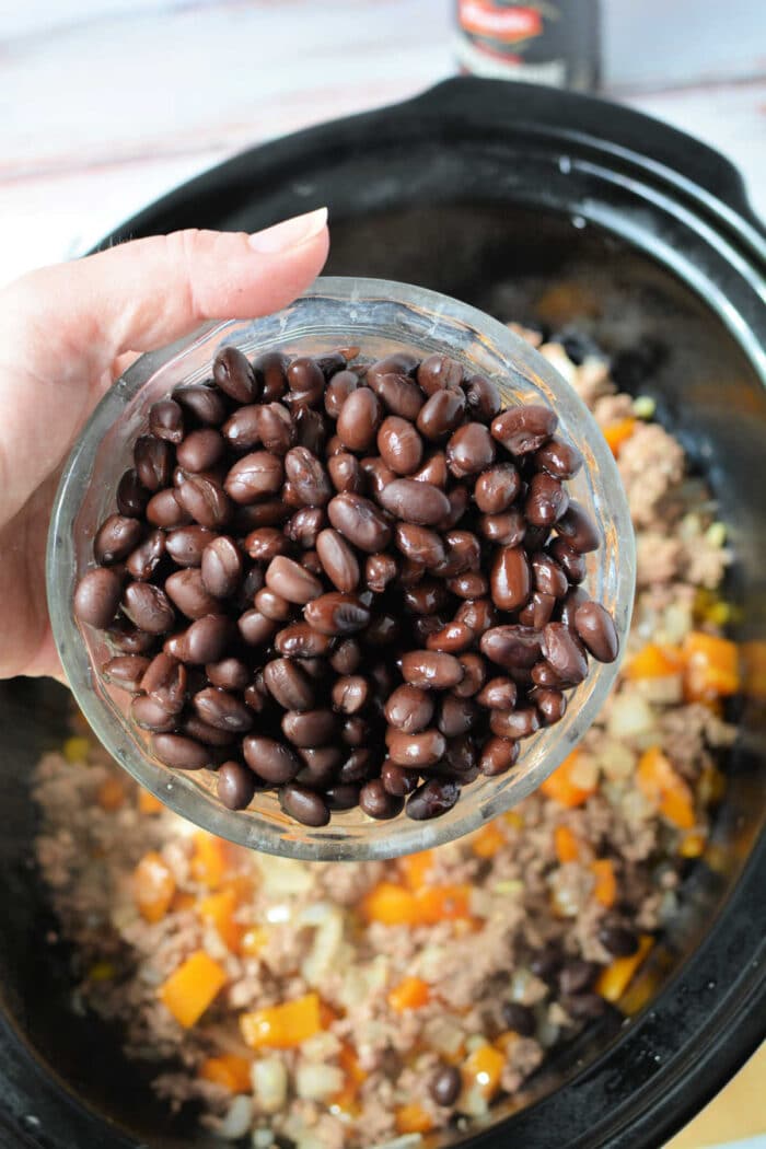 A hand holds a bowl of black beans over a Crockpot filled with ground meat and chopped vegetables, preparing a hearty Cowboy Casserole.