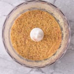 Seasoned breadcrumbs in a food processor bowl on a light marble countertop, viewed from above.