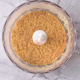 Seasoned breadcrumbs in a food processor bowl on a light marble countertop, viewed from above.