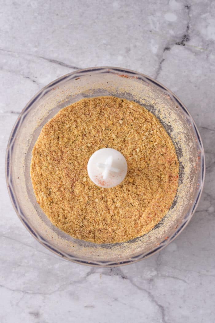 Food processor bowl with finely ground seasoned breadcrumbs on a light gray marble countertop.