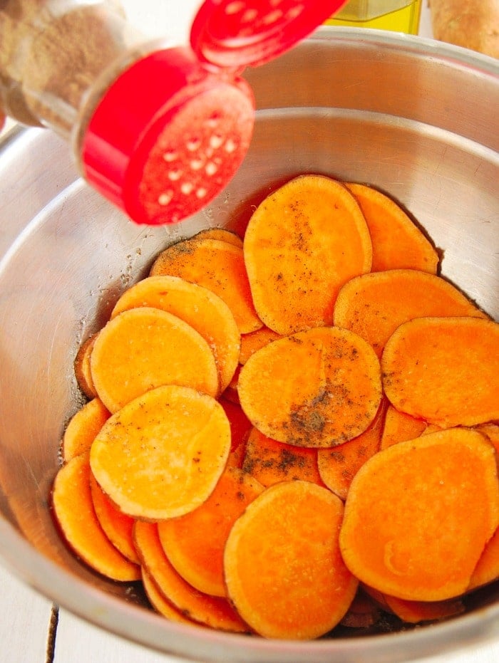 Sliced sweet potatoes in a metal bowl being sprinkled with seasoning from a red shaker, perfect for making sweet potato chips in the air fryer.