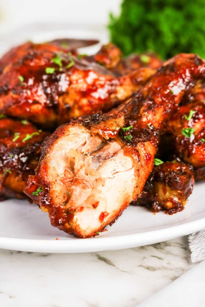 Oven baked BBQ chicken legs are served on a white plate, one with a bite taken out, and garnished with chopped herbs—a delicious addition to your collection of chicken recipes.