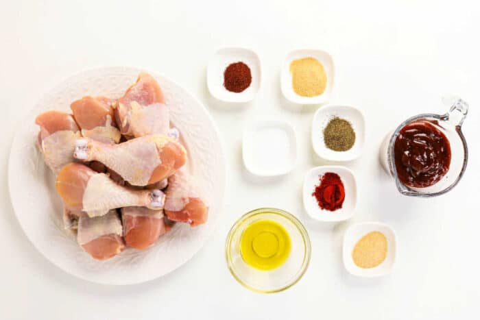 Raw chicken drumsticks on a plate with small bowls of spices, oil, salt, and barbecue sauce on a white background—perfect for your next oven baked BBQ chicken legs recipe.