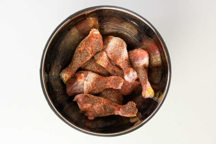 Raw chicken drumsticks coated with spices in a metal mixing bowl, viewed from above on a white background—perfect for preparing oven baked chicken or BBQ chicken legs.