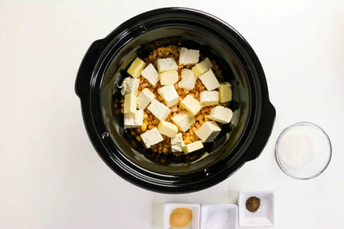 Crockpot Creamed Corn with corn and butter cubes, with bowls of sugar, garlic powder, pepper, and salt nearby.