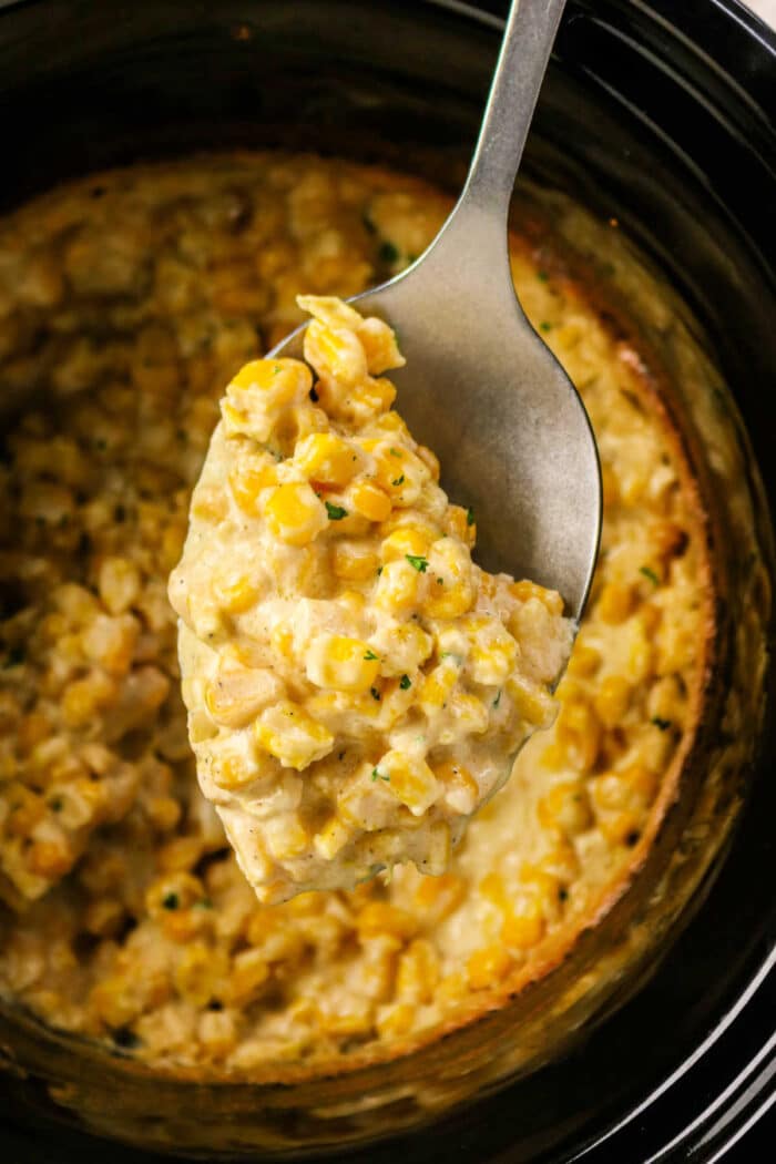 A spoon scooping creamy Crockpot Creamed Corn casserole from a slow cooker.