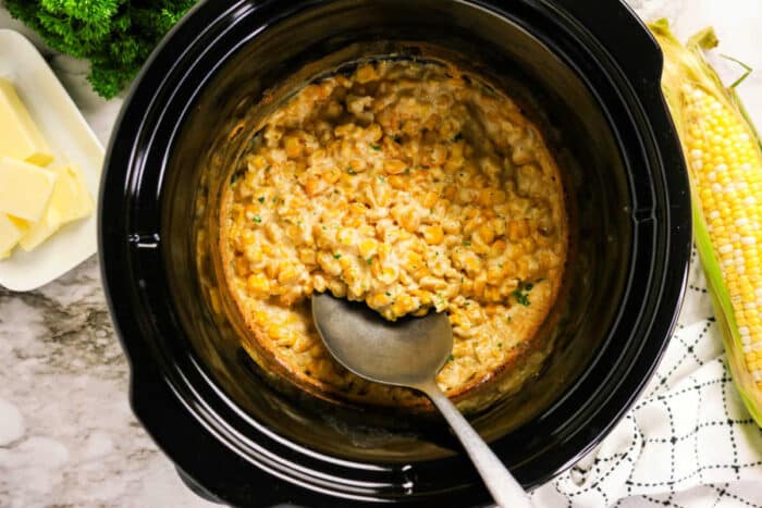 Crockpot Creamed Corn in a slow cooker with a serving spoon, fresh corn, butter, and herbs on the side.