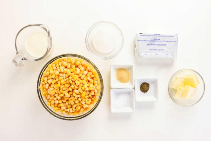 Bowls of corn, butter, seasonings, cream, and sugar on a white surface, ready for cooking the perfect Crockpot Creamed Corn.