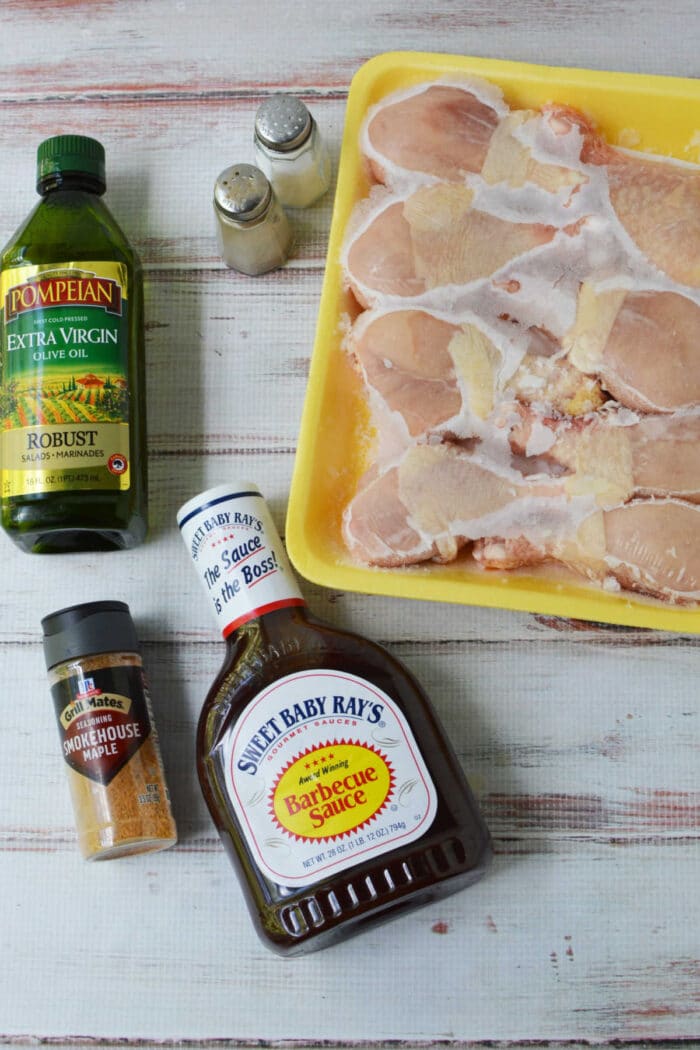 Raw chicken legs in a yellow tray with olive oil, barbecue sauce, seasoning, and salt and pepper shakers—everything you need to prepare delicious baked chicken legs or use frozen chicken legs for a quick and easy meal.