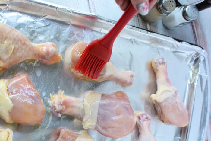 A hand brushes raw frozen chicken legs with oil on a foil-lined baking sheet, salt and pepper nearby, preparing them perfectly for baked frozen chicken.