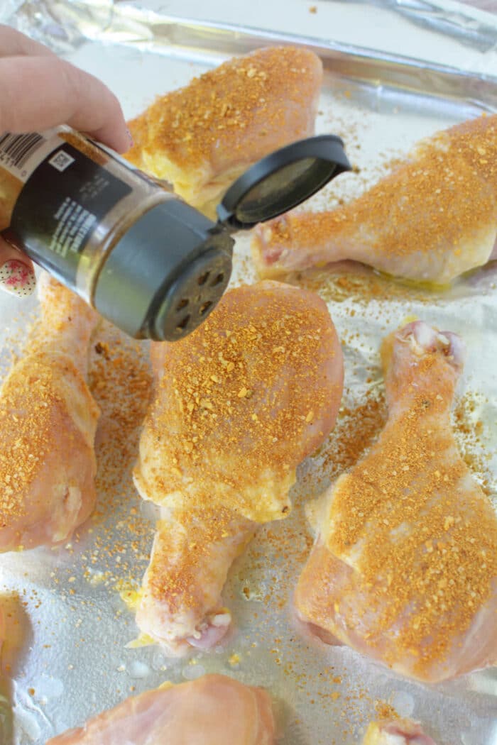 A hand sprinkles seasoning onto raw chicken drumsticks on a foil-lined baking sheet, preparing them to become flavorful baked chicken legs.
