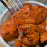A fork holds a glazed teriyaki salmon bite over a bowl filled with more teriyaki salmon bites.