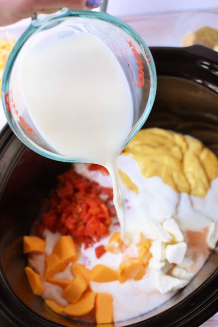A measuring cup pours milk into a slow cooker filled with cheese, diced tomatoes, and all the delicious ingredients needed for Crockpot Chicken Spaghetti.