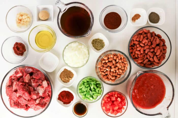 Bowls of raw beef, beans, diced vegetables, spices, and liquids arranged on a white surface, viewed from above—ingredients perfectly suited for a hearty Chuck Roast Chili.