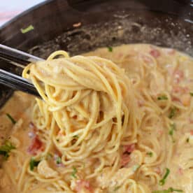 Tongs lifting creamy chicken spaghetti with tomatoes and herbs from a black bowl, fresh from the crockpot.