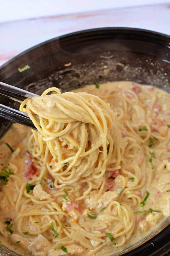 Tongs lifting creamy chicken spaghetti with tomatoes and herbs from a black bowl, fresh from the crockpot.