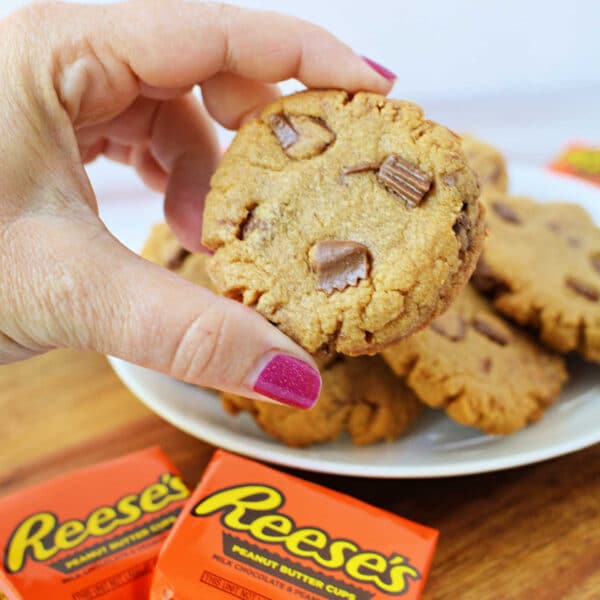 A hand with pink nail polish holds a delicious peanut butter cookie with Reeses pieces over a plate of freshly-baked Peanut Butter Cookies.