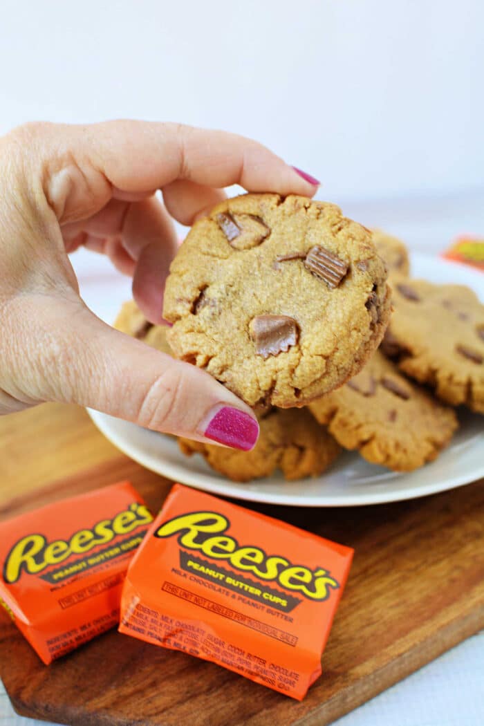 A hand with pink nail polish holds a delicious peanut butter cookie with Reeses pieces over a plate of freshly-baked Peanut Butter Cookies.
