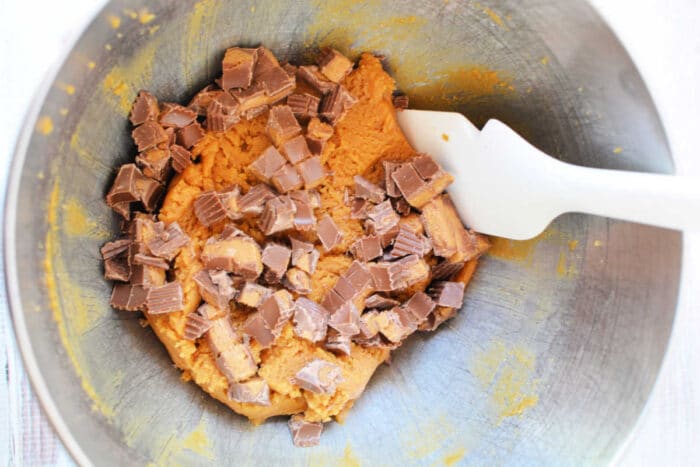 Cookie dough with chopped chocolate candy pieces and bits of Peanut Butter Cups, stirred together with a white spatula in a metal mixing bowl—perfect for making the ultimate Peanut Butter Cookies.