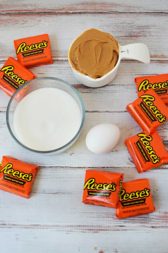 A bowl of sugar, creamy peanut butter, an egg, and several Reese’s peanut butter cups are arranged on a white surface—perfect for making irresistible peanut butter cookies.