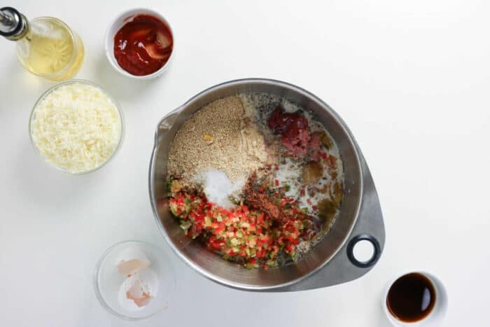 Metal bowl with eggs, breadcrumbs, diced peppers, and seasonings&mdash;perfect for mixing up a delicious Philly Cheesesteak Meatloaf. Cheese, ketchup, and sauces are arranged in small bowls nearby to enhance the classic flavors of this meatloaf-inspired dish.