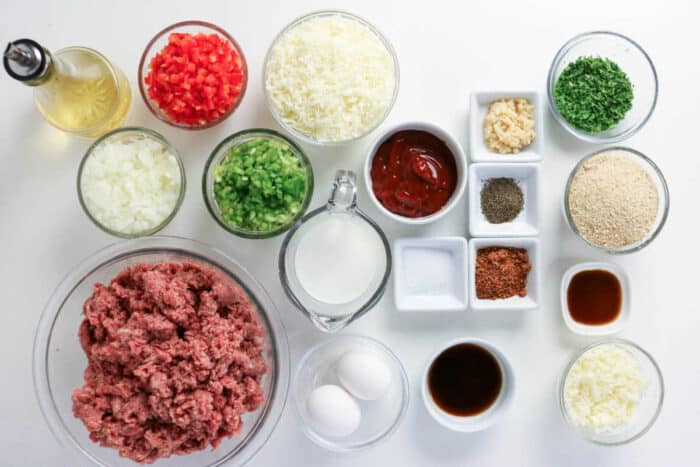 Ground meat, eggs, vegetables, seasonings, cheese, breadcrumbs, milk, and sauces arranged on a white surface&mdash;everything you need to craft a delicious Philly Cheesesteak Meatloaf bursting with classic flavors.