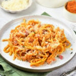 Plate of pasta with red sauce and grated cheese, featuring a delicious twist of Pumpkin Spaghetti, with small bowls of cheese, diced ham, and sauce in the background.
