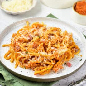 Plate of pasta with red sauce and grated cheese, featuring a delicious twist of Pumpkin Spaghetti, with small bowls of cheese, diced ham, and sauce in the background.