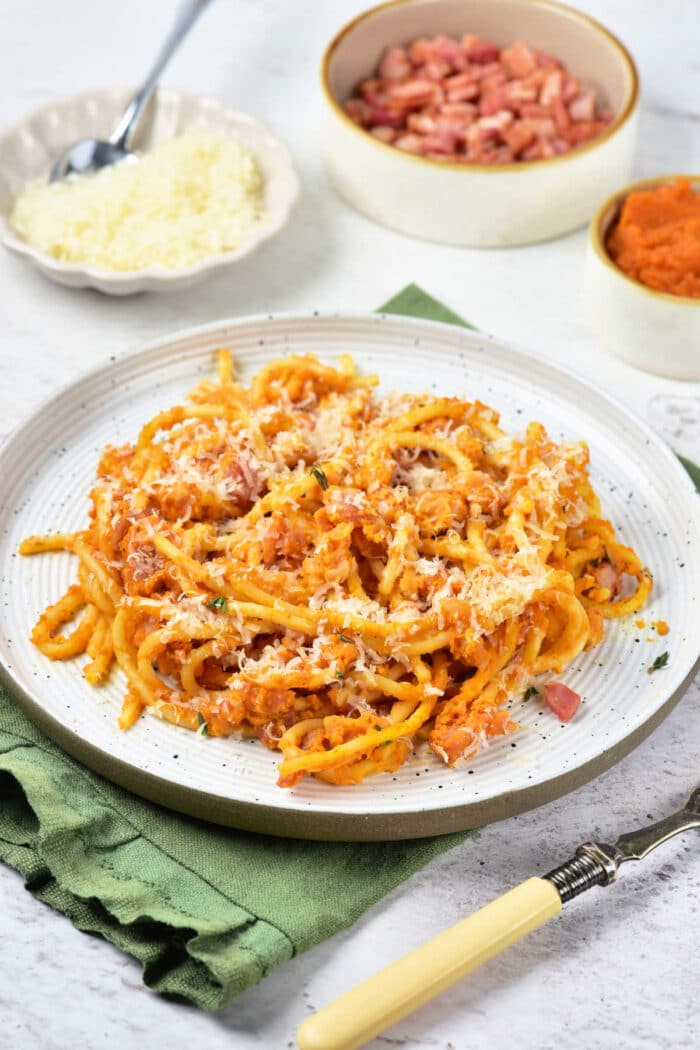 Plate of pasta with red sauce and grated cheese, featuring a delicious twist of Pumpkin Spaghetti, with small bowls of cheese, diced ham, and sauce in the background.