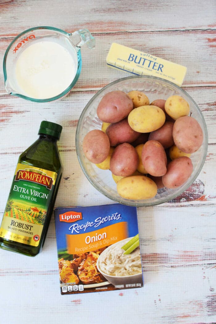 Bowl of potatoes, butter, milk, olive oil, and onion soup mix on a white wooden table—perfect ingredients for trying delicious Slow Cooker Potatoes or adding to your favorite slow cooker recipes.