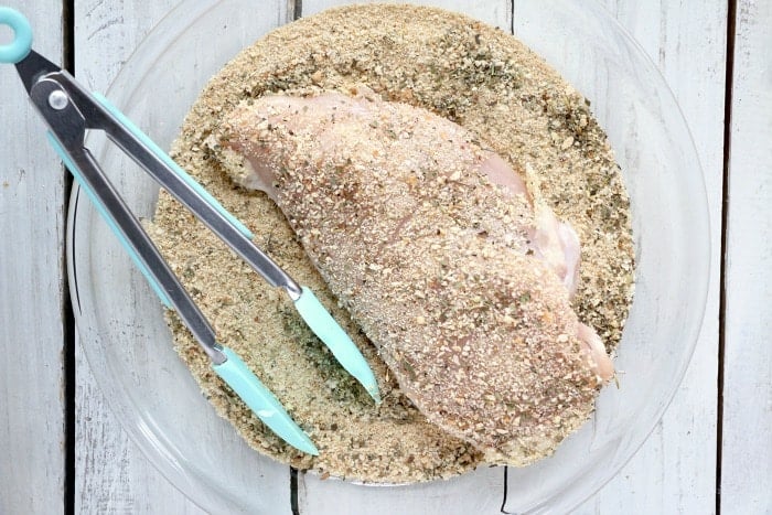 Raw chicken breast being coated in seasoned breadcrumbs with tongs in a glass dish on a white wooden surface, preparing for a delicious slow cooker chicken parmesan.
