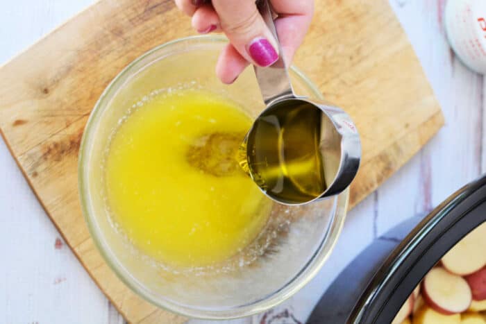 Hand pours oil from a metal cup into a bowl of melted butter on a wooden cutting board, creating the perfect base for delicious slow cooker potatoes or other potato recipes.