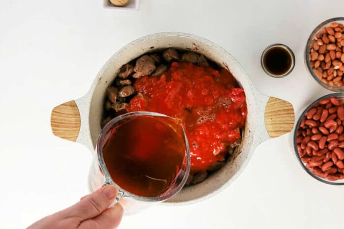 A hand pours broth into a pot with beef and tomatoes, surrounded by beans and sauce on a white table, creating the perfect base for a hearty beef chili recipe.