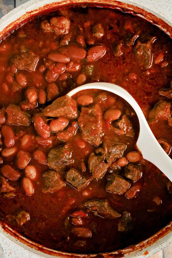 A white ladle scoops rich Chuck Roast Chili, brimming with beef and beans, from a pot filled with thick, red, hearty stew.