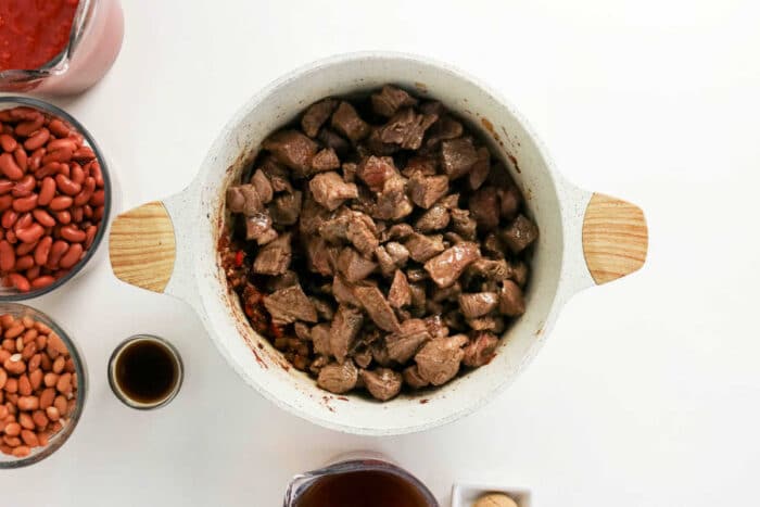 Overhead view of cubed beef in a white pot, perfect for making Chuck Roast Chili, surrounded by beans, sauce, and seasonings on a white surface.