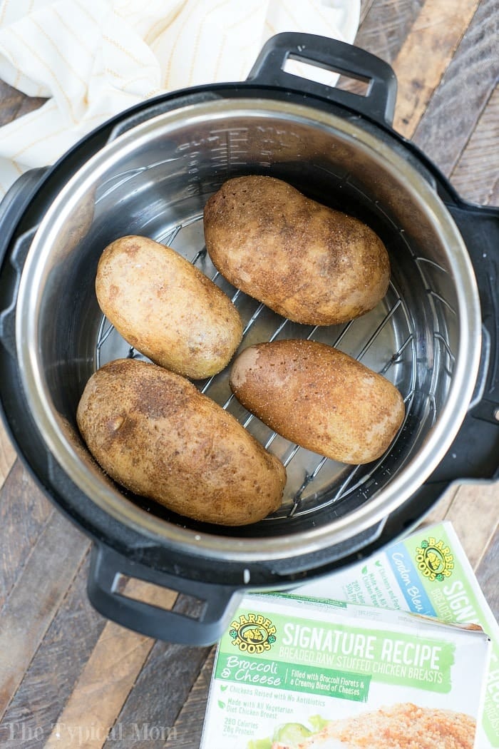 Pressure cooker baked potatoes and stuffed chicken are a great duo! Instant Pot dinner success again. #pressurecooker #instantpot #bakedpotatoes #potatoes #sidedish #instantpotrecipes #thetypicalmom #ninjafoodi Four whole russet potatoes are nestled on a metal rack inside an Instant Pot, ready to become delicious instant pot baked potatoes, with a food box nearby on a wood table.