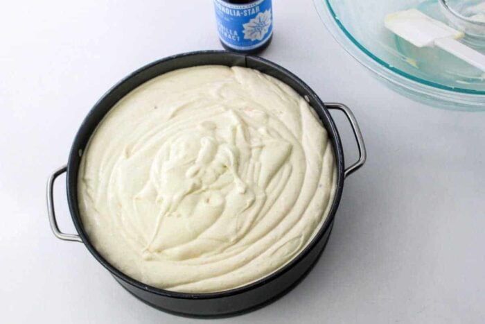 Round cake pan filled with cheesecake batter on a white surface next to a spatula and vanilla extract, ready for your favorite Philadelphia Cream Cheese Cheesecake Recipe.