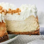 A slice of cheesecake made from a classic Philadelphia Cream Cheese Cheesecake Recipe, topped with whipped cream and cookie crumbs, is being lifted from a plate.