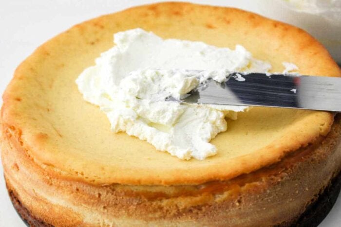 Spreading whipped cream on top of a plain cheesecake made from the classic Philadelphia Cream Cheese Cheesecake Recipe with a metal spatula.