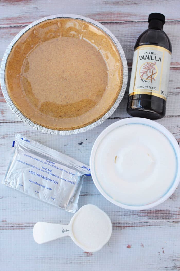 An empty pie crust, vanilla extract, cream cheese, and a scoop for a 3 ingredient cheesecake.
