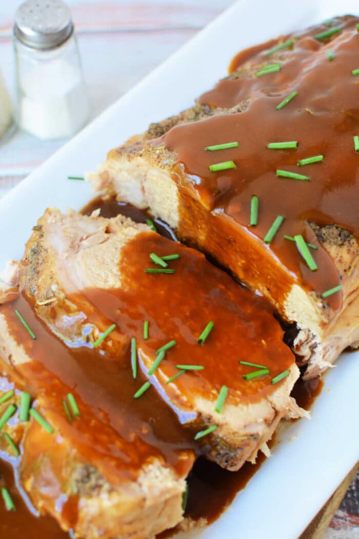 Sliced oven-baked pork loin covered in brown gravy and garnished with chopped chives on a white platter.
