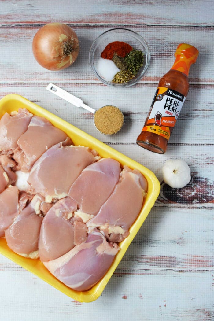 Raw chicken thighs, a bottle of Nando’s sauce, aromatics, and spices for a slow cooker peri-peri chicken recipe.