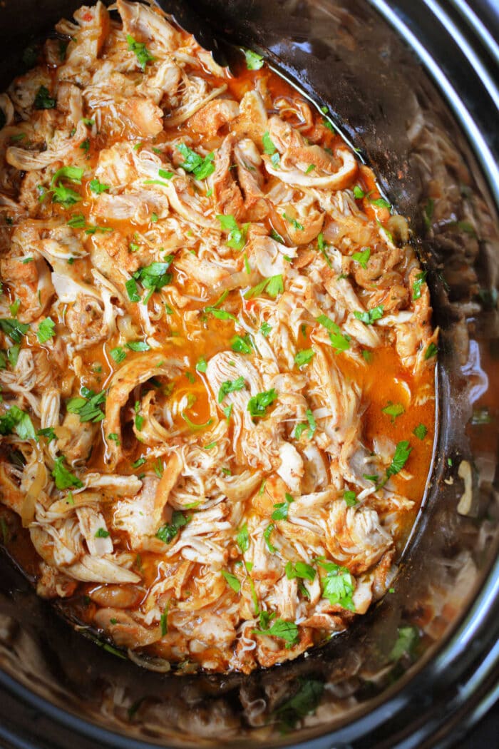 Shredded peri peri chicken in a flavorful red sauce, garnished with fresh parsley inside a slow cooker.